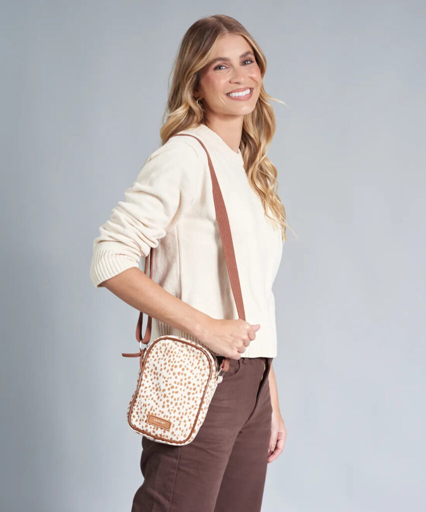 Mulher sorrindo com uma bolsa transversal pequena de estampa poá em tons de bege e marrom, com alça marrom, usando suéter bege e calça marrom, sobre fundo cinza neutro.