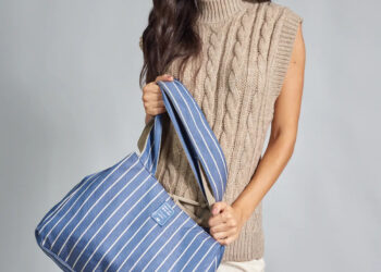 Mulher segurando uma bolsa tote azul com listras verticais brancas e alças largas, usando blusa de tricô sem mangas em tom bege e calça clara, em fundo cinza neutro.