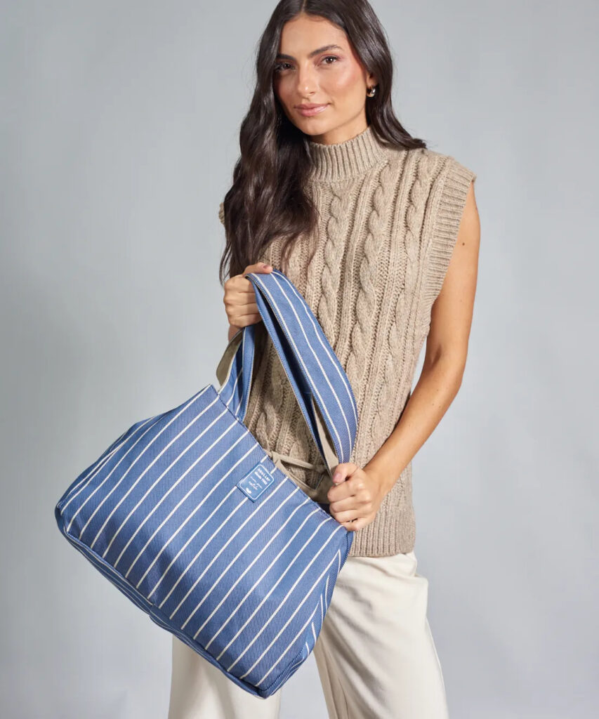 Mulher segurando uma bolsa tote azul com listras verticais brancas e alças largas, usando blusa de tricô sem mangas em tom bege e calça clara, em fundo cinza neutro.