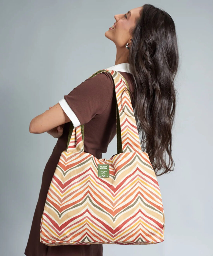 Mulher usando uma bolsa tote estampada com padrão chevron em tons de coral, bege, amarelo e verde, com alças largas, vestindo vestido marrom de gola polo, em fundo cinza neutro.