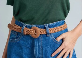 Mulher usando um cinto marrom trançado com fivela redonda, combinado com calça jeans azul e blusa verde, em fundo cinza neutro.